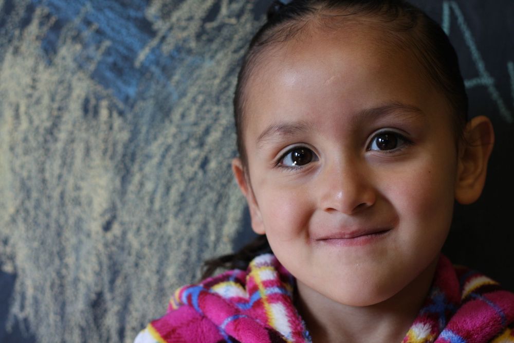 A child wearing a pink jacket smiles warmly against a dark background with white chalk marks.