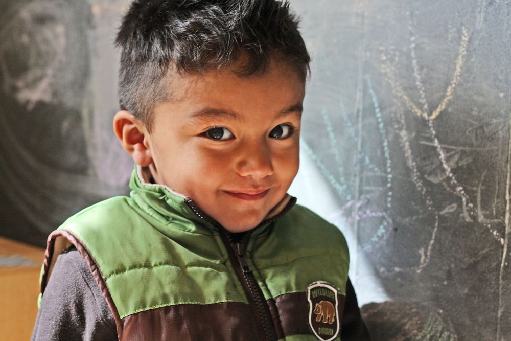 A child wearing a brown and green vest smiles at the camera while standing in front of a chalkboard.