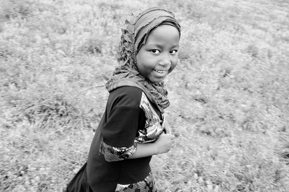 Black and white portrait showing a joyful young person in traditional headscarf smiling at camera in an outdoor setting.