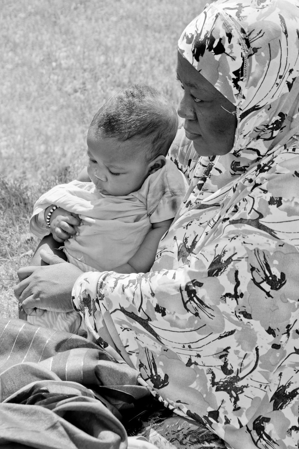 A tender black and white portrait of a figure in a floral headscarf sitting outdoors with a small baby.