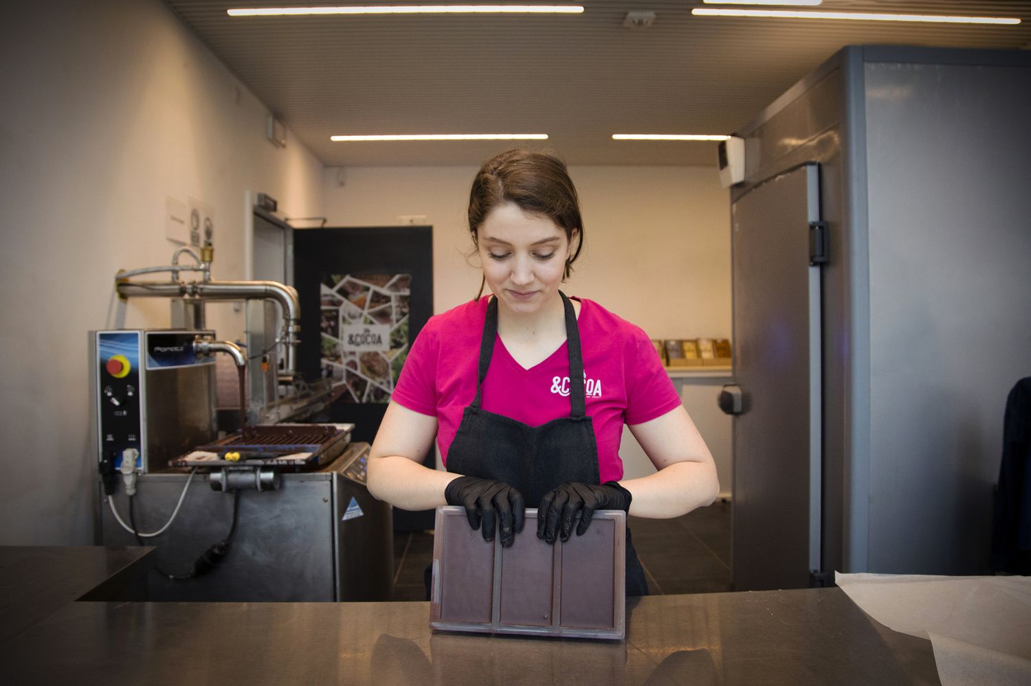 Un artisan chocolatier portant une chemise rose et des gants noirs manipule des moules de tablettes de chocolat