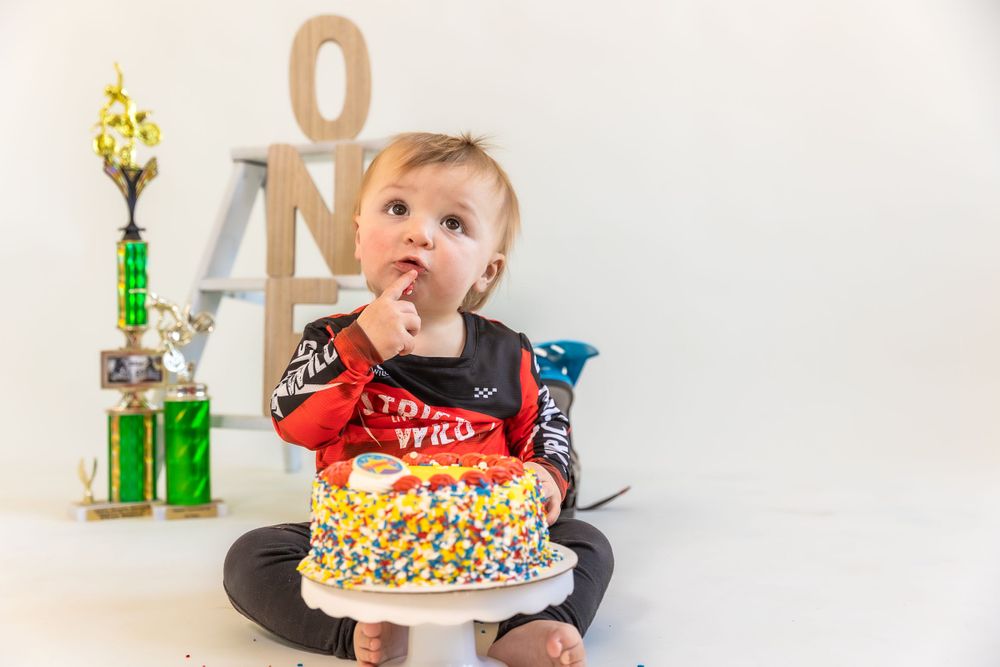 A young toddler in a red vest sits on a floor in front of a first birthday cake with a trophy visible in the background.