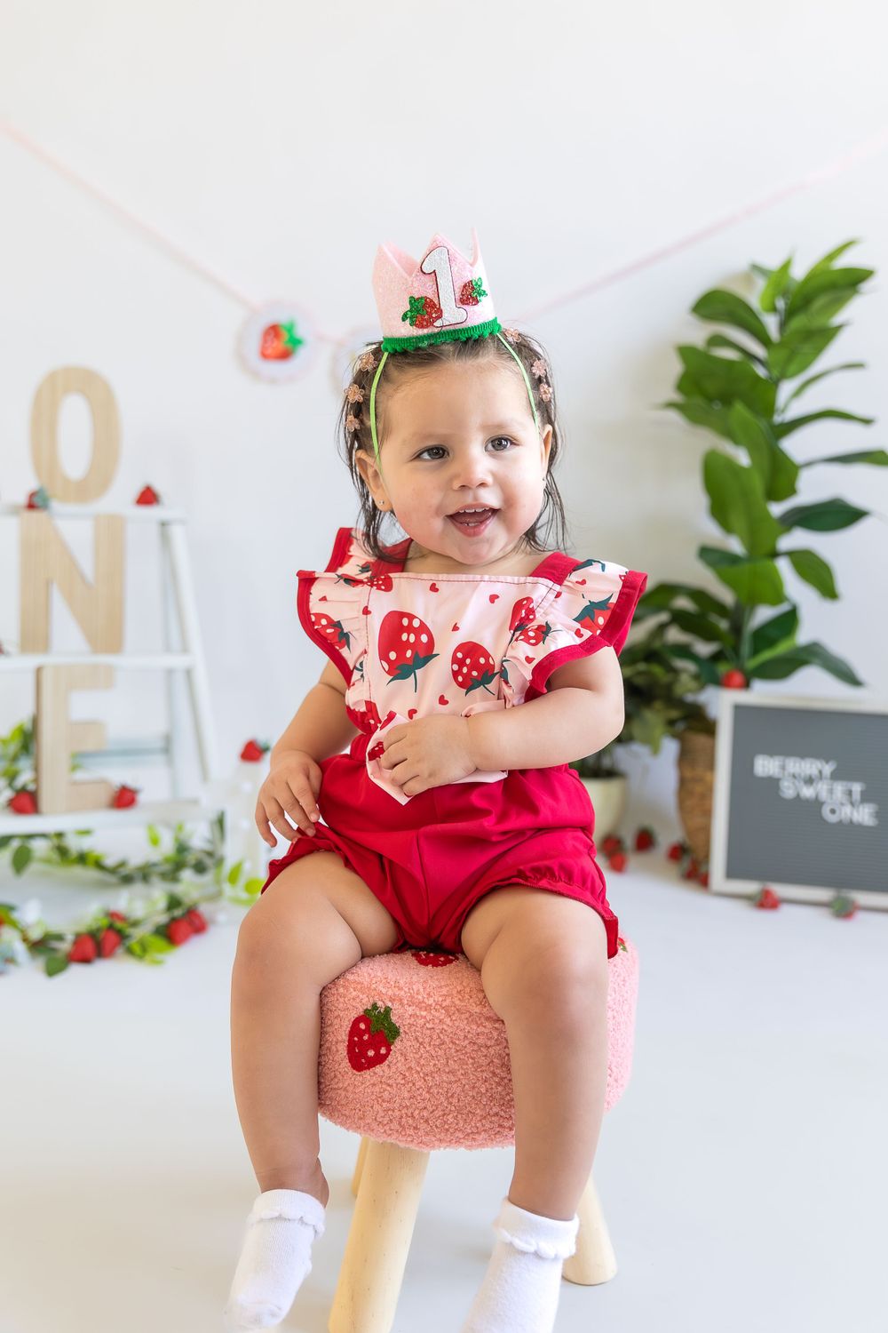 Child in strawberry-themed outfit sits on pink mushroom stool with decorative party setup in background.
