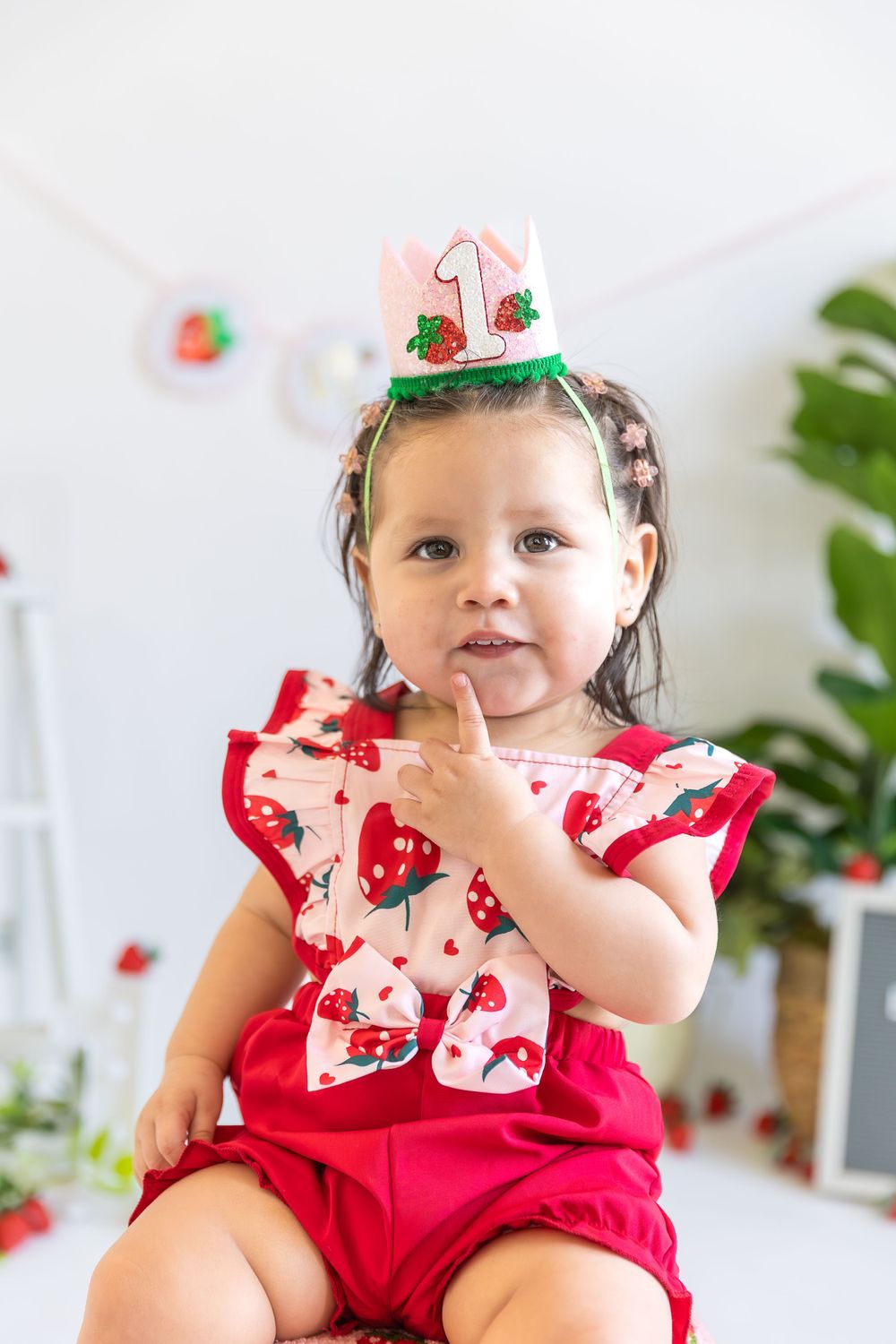 Toddler wearing red romper with strawberry print and miniature birthday hat celebrating first birthday.