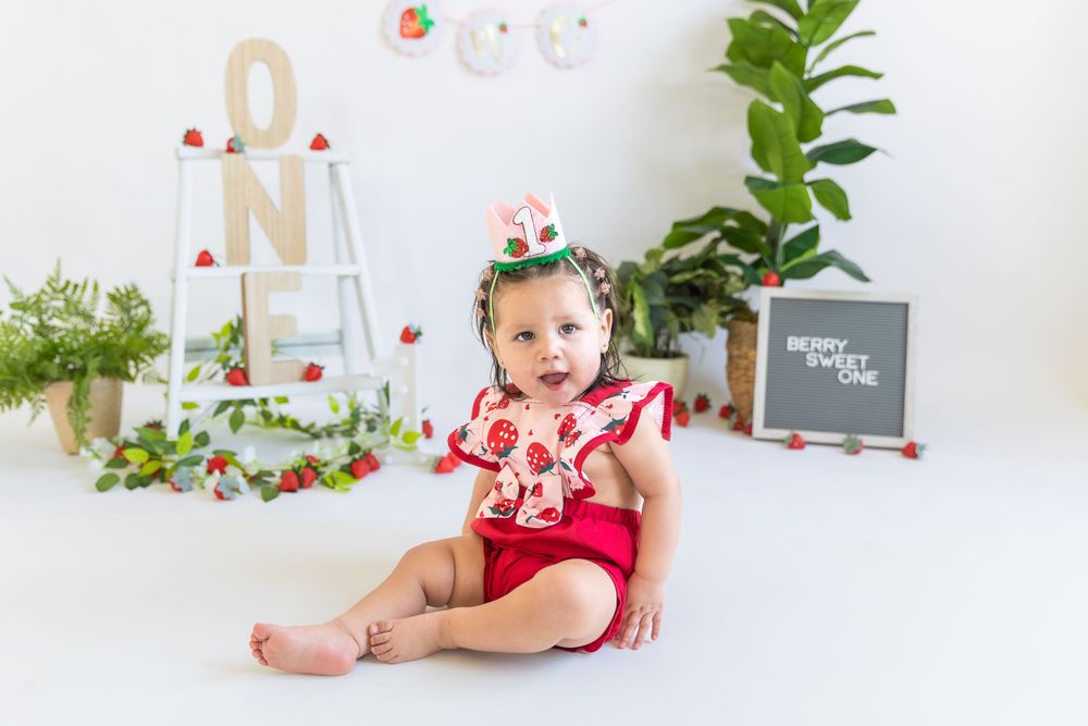 A child in red and floral outfit sits on white floor with decorative ladder and chalkboard in the background.
