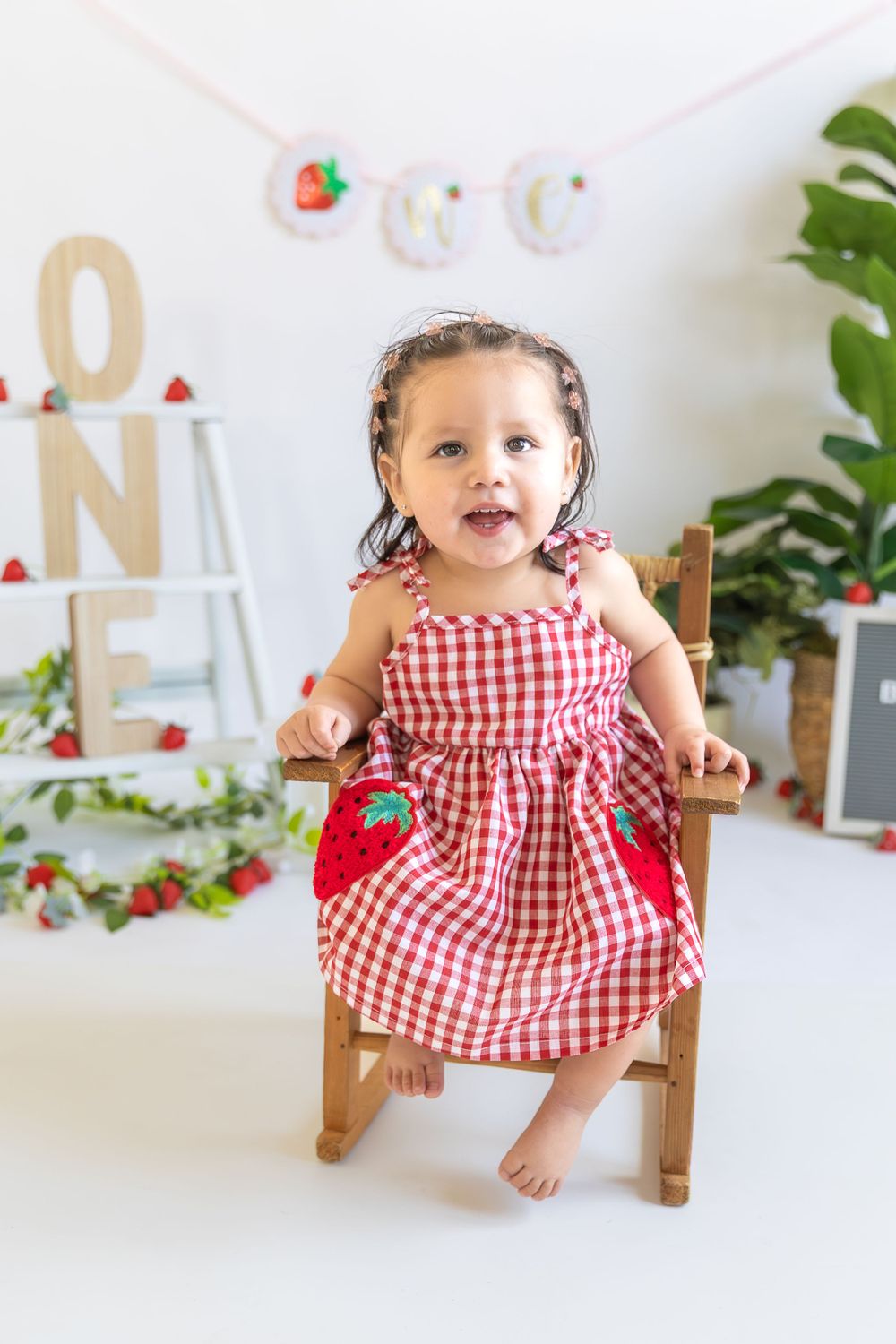 Small child wearing red and white gingham dress sits in wooden chair against white backdrop with festive decorations.