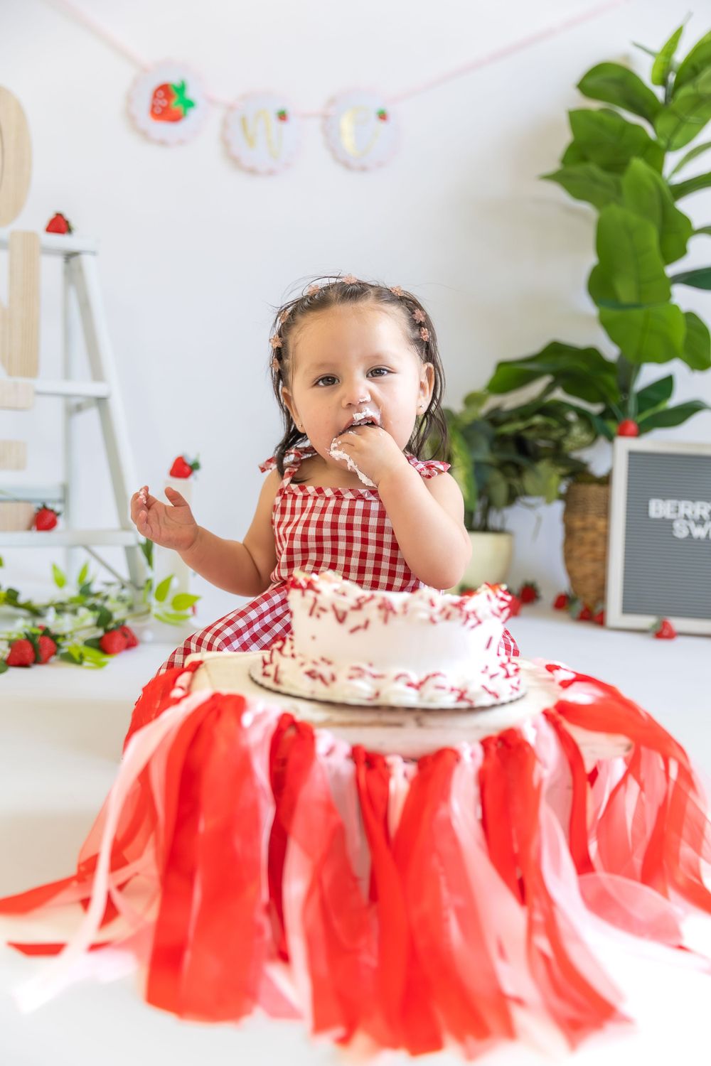 Festive birthday celebration setup with red gingham dress, red tulle tablecloth and white cake with strawberry decorations.