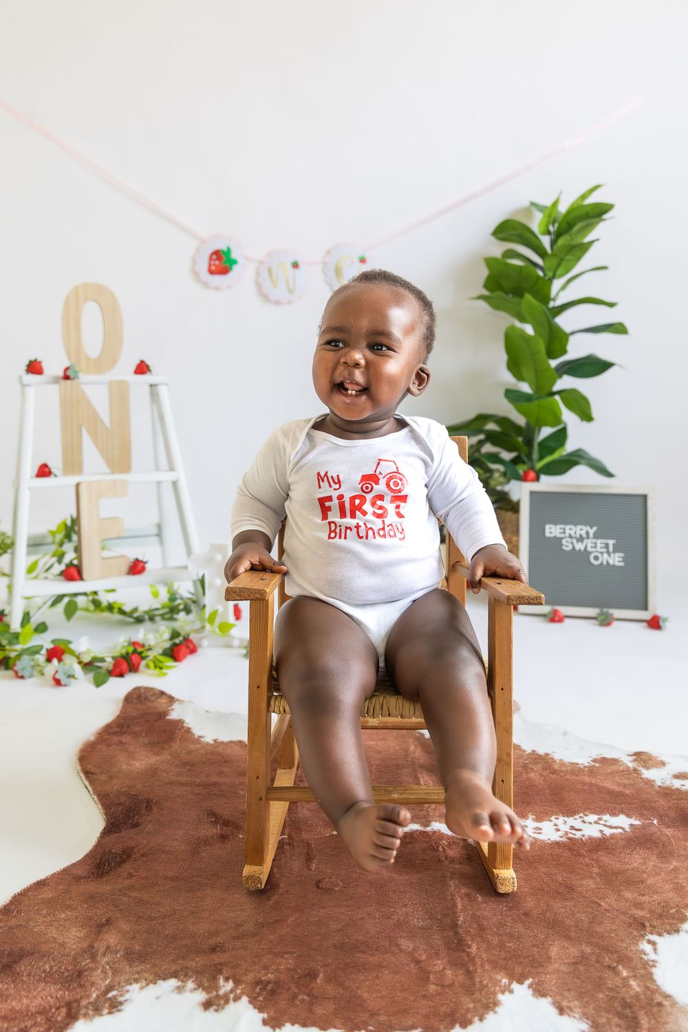 Baby sitting on wooden rocking chair against white wall with holiday decor and house plant.