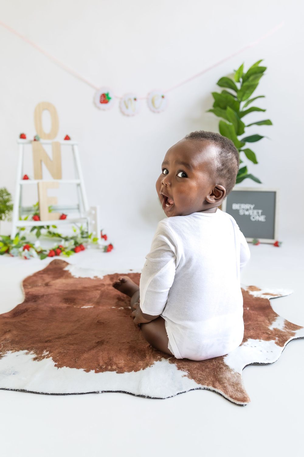 Baby in white onesie sits on brown and white cowhide rug with decorative display in background.