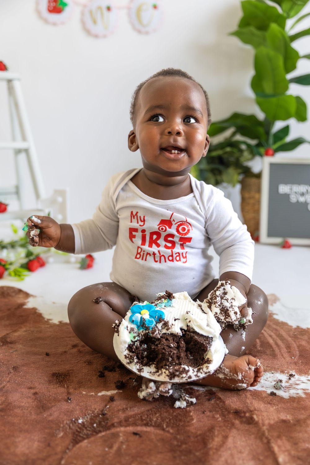 Baby in "My First Birthday" shirt sits with smashed chocolate cake during cake smash photo session.