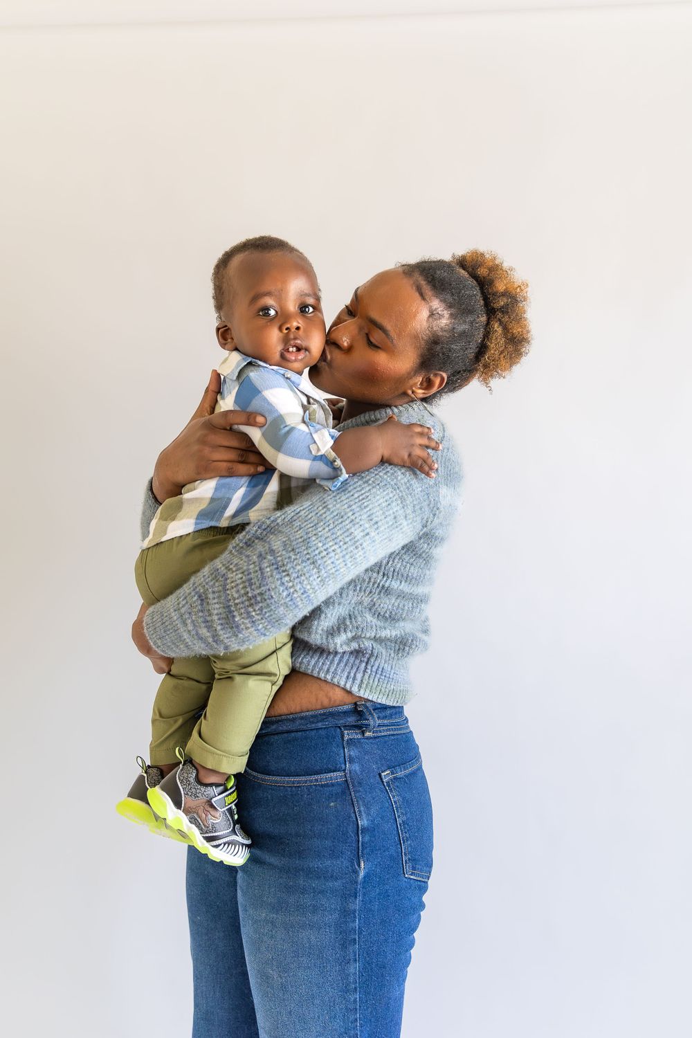 A parent in a gray sweater and jeans lifts and embraces a young child wearing a striped shirt and green pants.