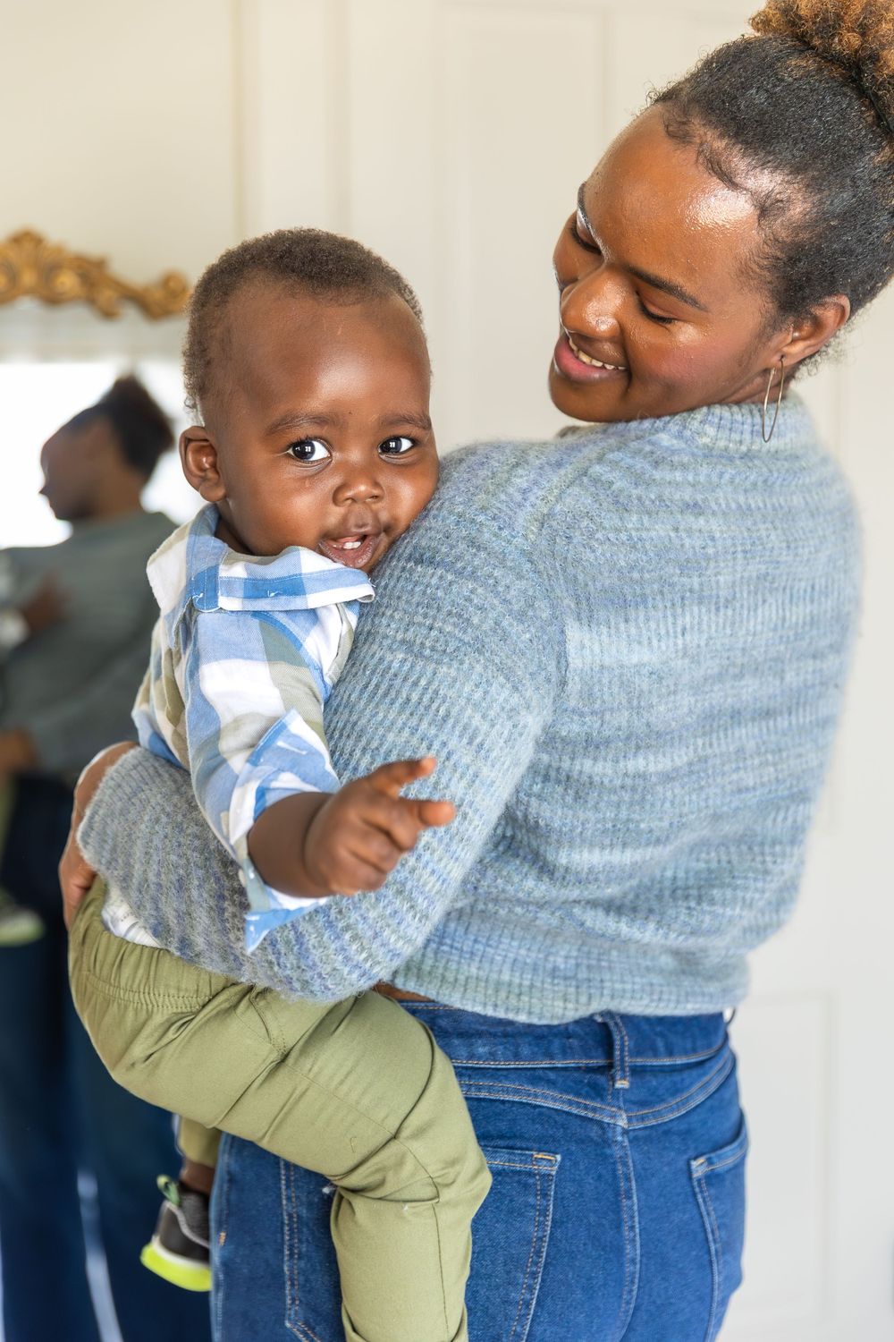 A parent in a gray sweater holds a smiling baby wearing a blue and white plaid shirt and olive pants.