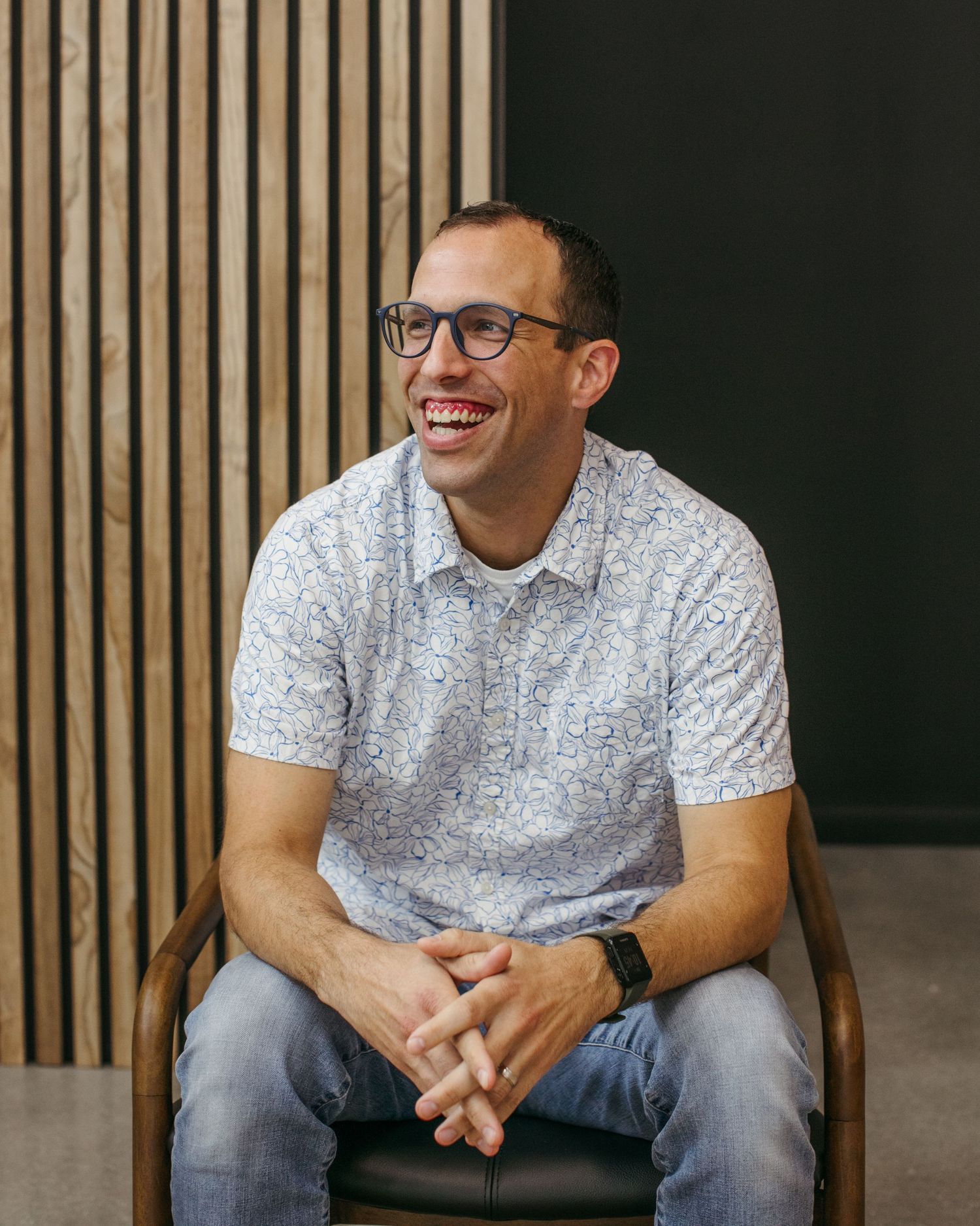A person in glasses and a white patterned shirt sits smiling in front of a wooden slat wall.