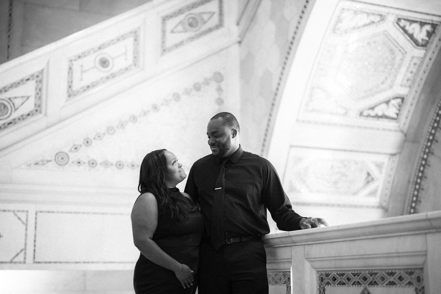 Romantic black and white photo of a couple sharing a moment on grand staircase with ornate decorative ceiling details.