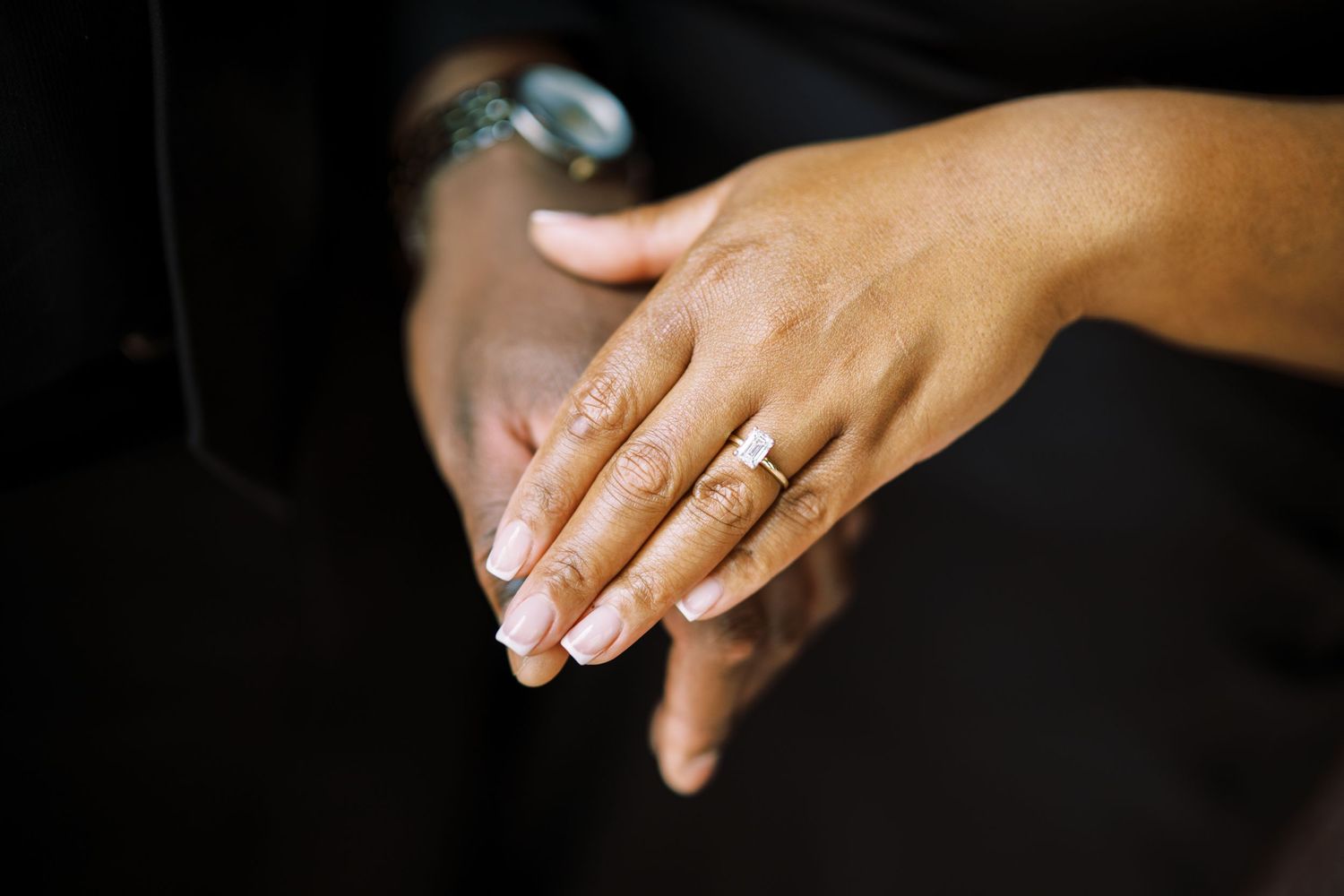 Close-up of hands showing off an elegant engagement ring with French manicured nails against a dark background.