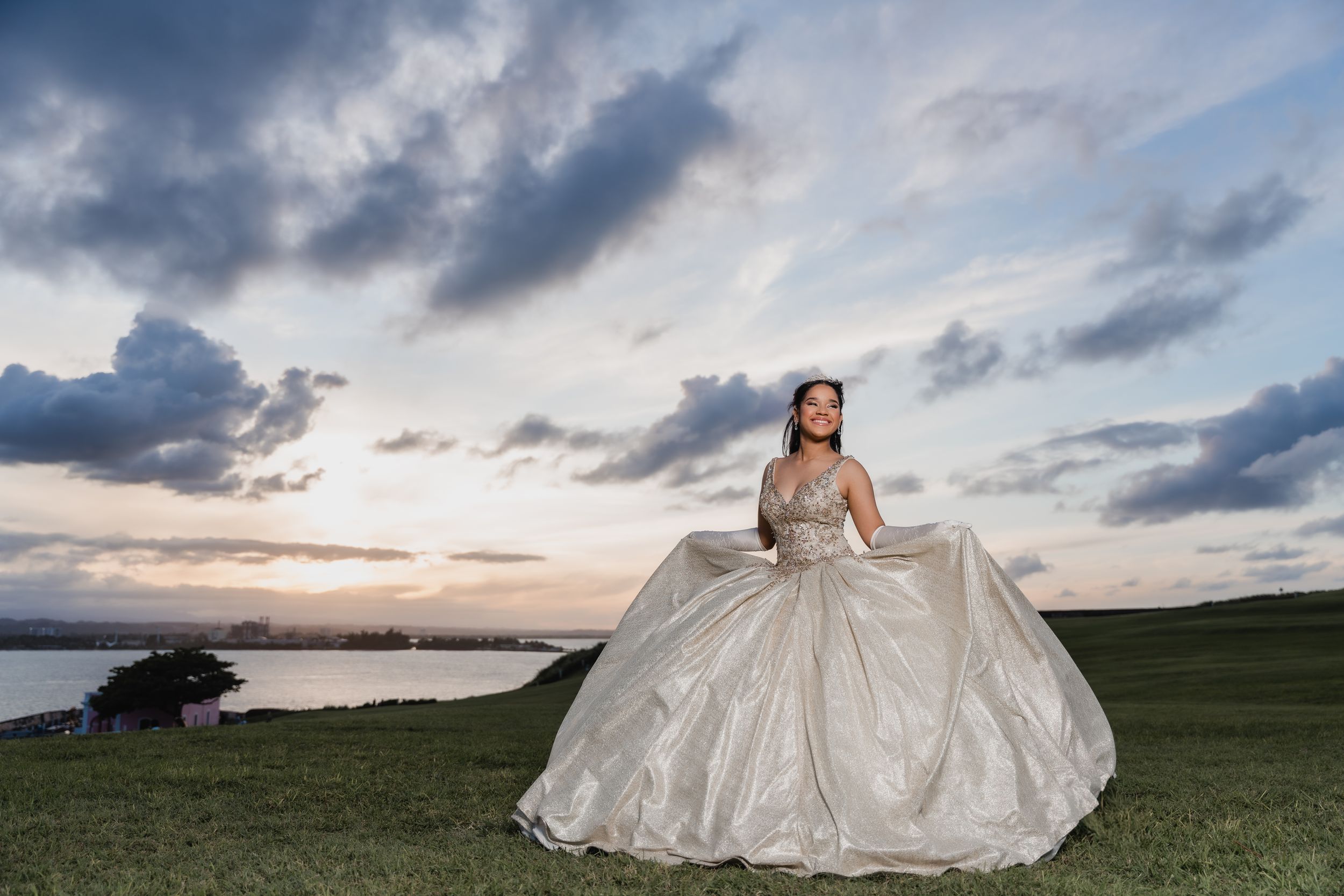 A person in a sparkly white ballgown poses on a grassy hill at sunset with dramatic clouds in the sky.