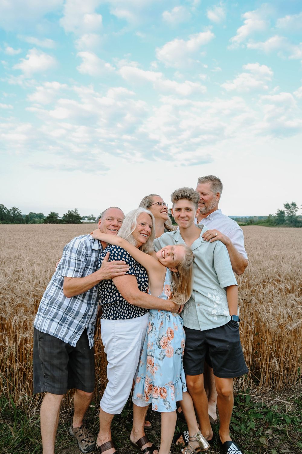 A happy group wearing casual attire poses together and laughs in front of a golden wheat field on a sunny day.