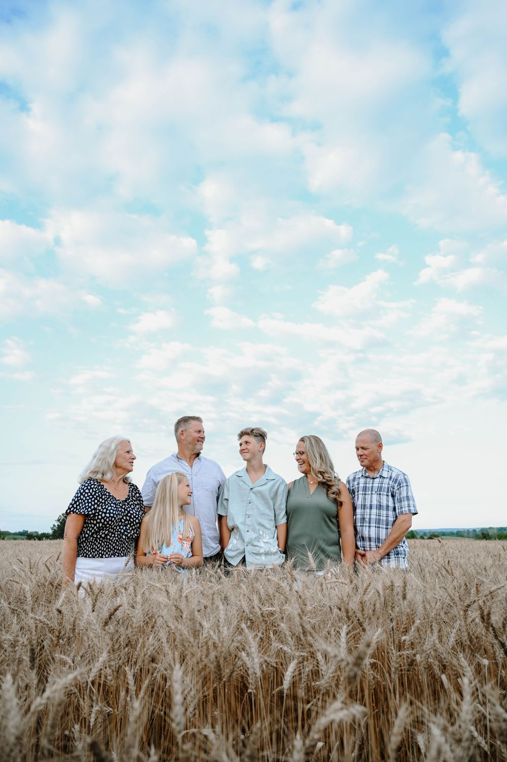 A family stands together in a golden wheat field under a blue cloudy sky during a summer day.