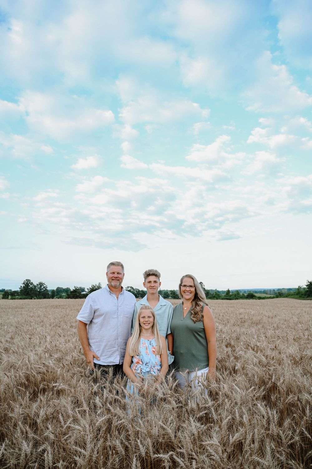 A family stands together in a golden wheat field beneath a bright blue summer sky with wispy clouds.