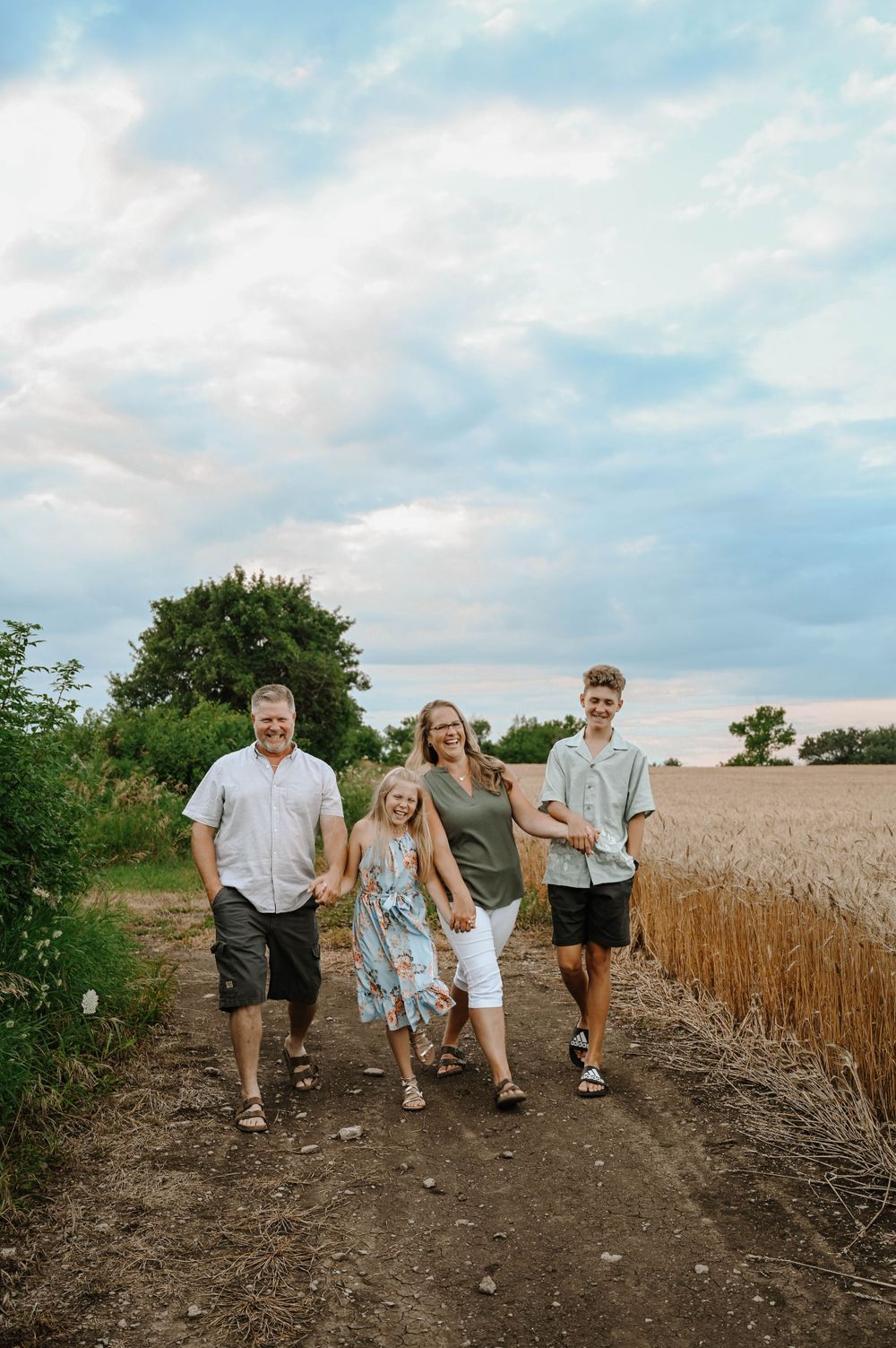 A family walks together on a dirt path between a wheat field and green shrubs on a summer day with cloudy skies.