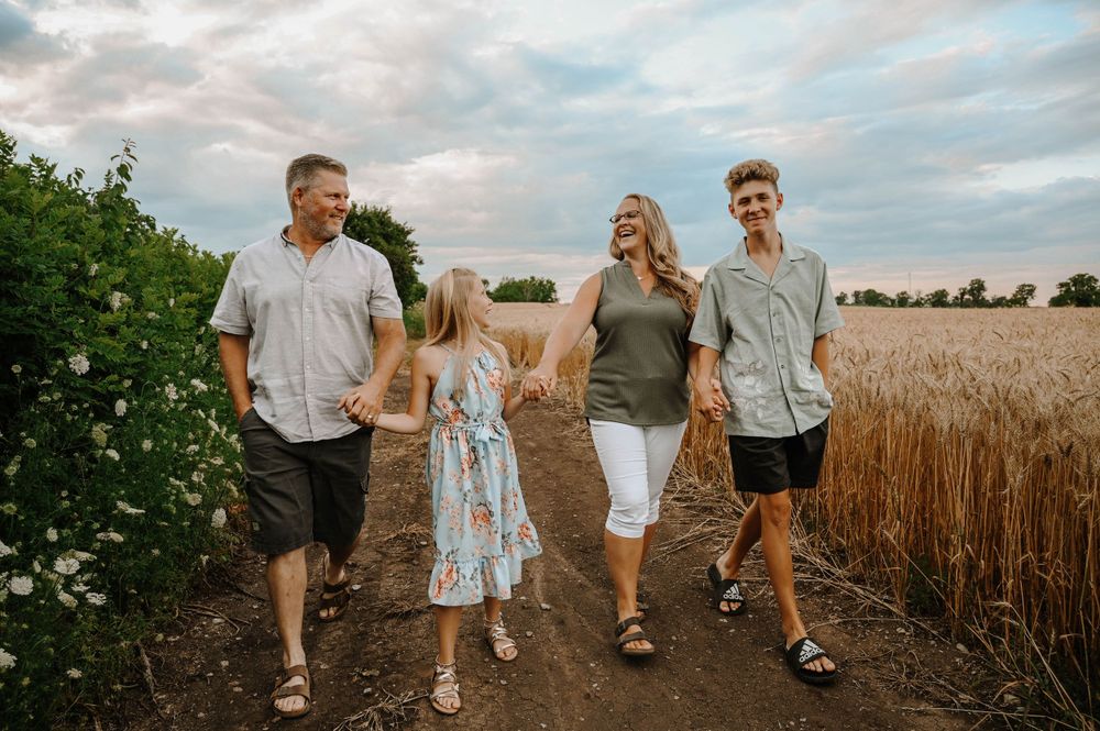 A family walks together on a dirt path between a wheat field and flowering bushes at sunset.