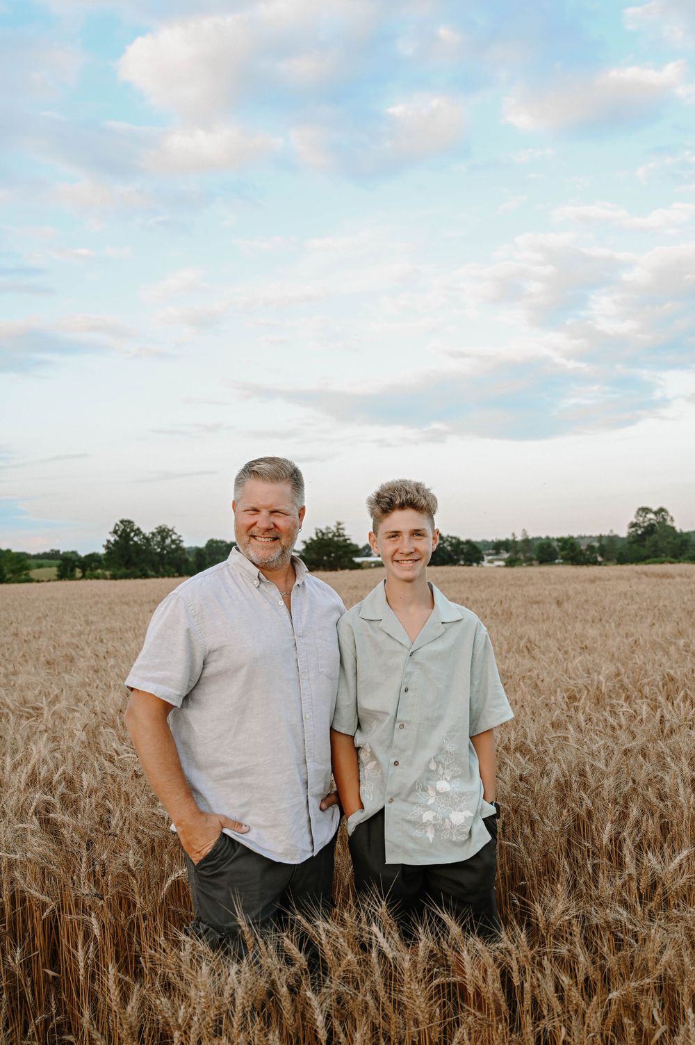 Two people stand together smiling in a golden wheat field on a summer day with light clouds in the blue sky.