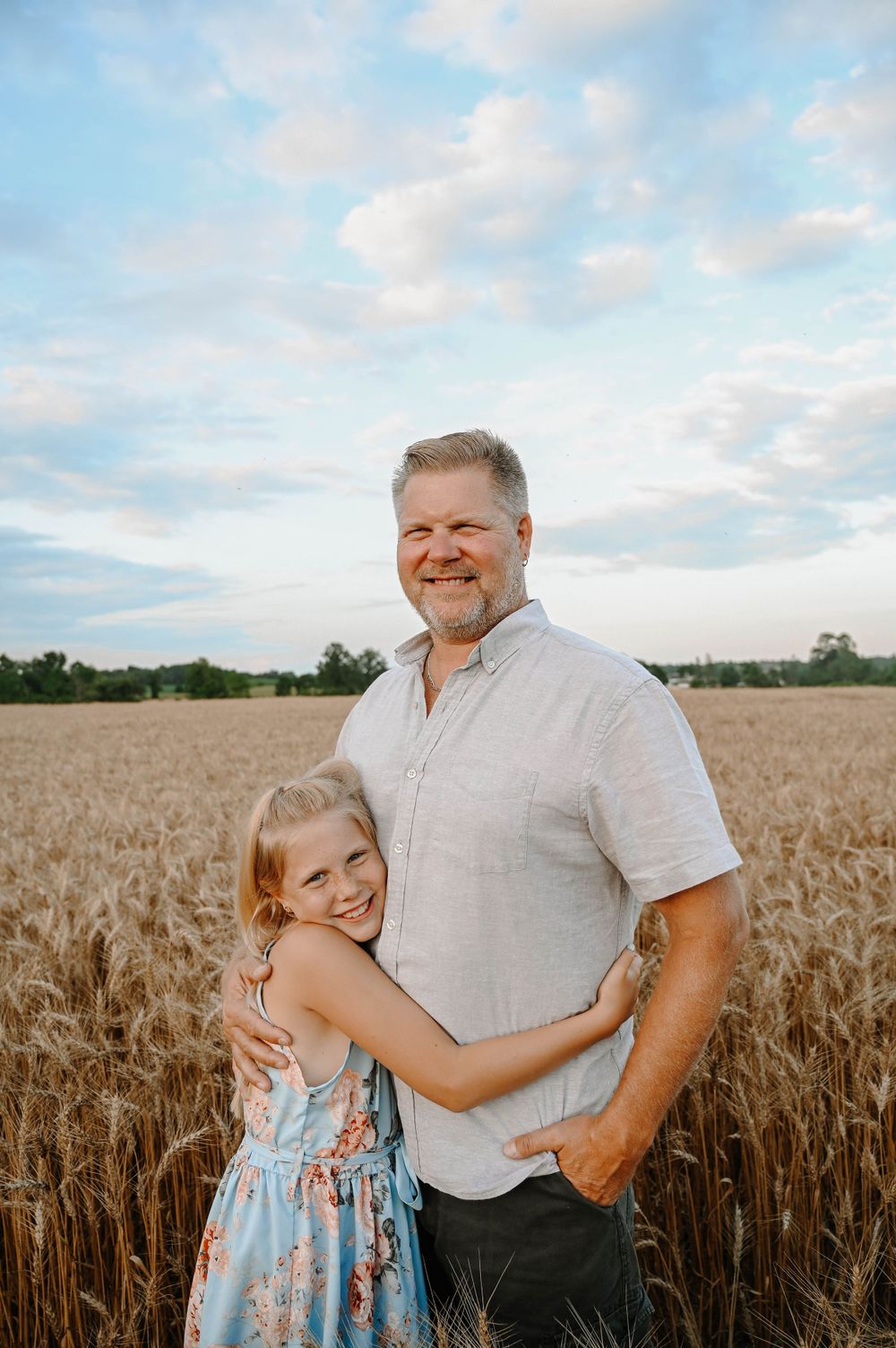 A loving moment captured in a golden wheat field during sunset with a blue sky and wispy clouds in the background.