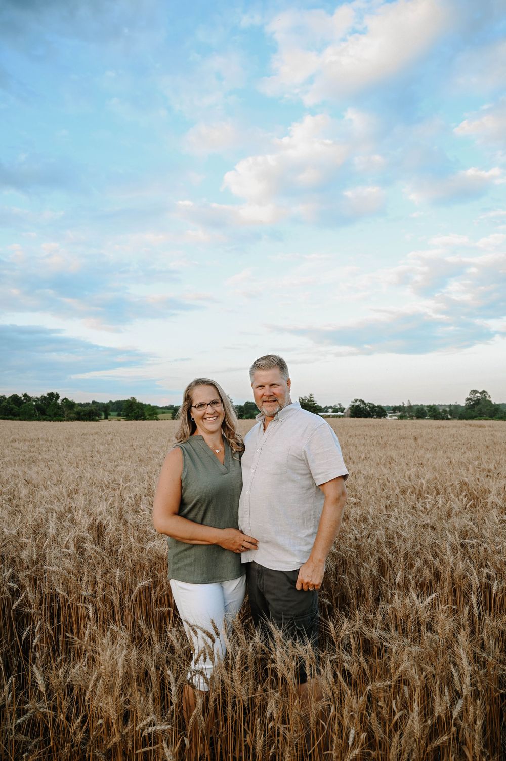 A couple stands together in a golden wheat field under a bright blue summer sky with wispy clouds.