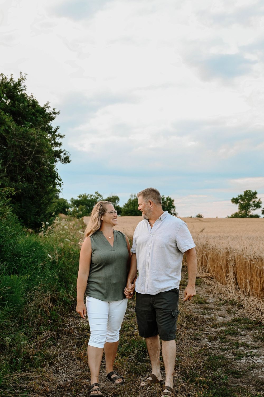 A couple walks together through a wheat field on a summer day with blue skies and trees in the background.