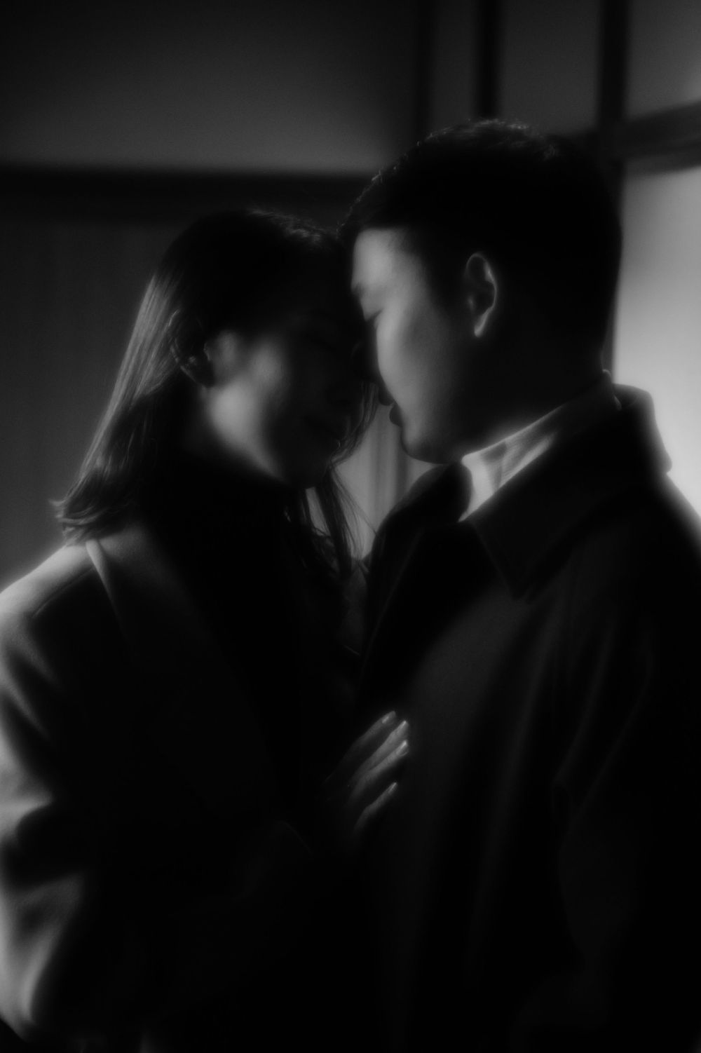A softly blurred black and white image of a couple standing close together in dim light.