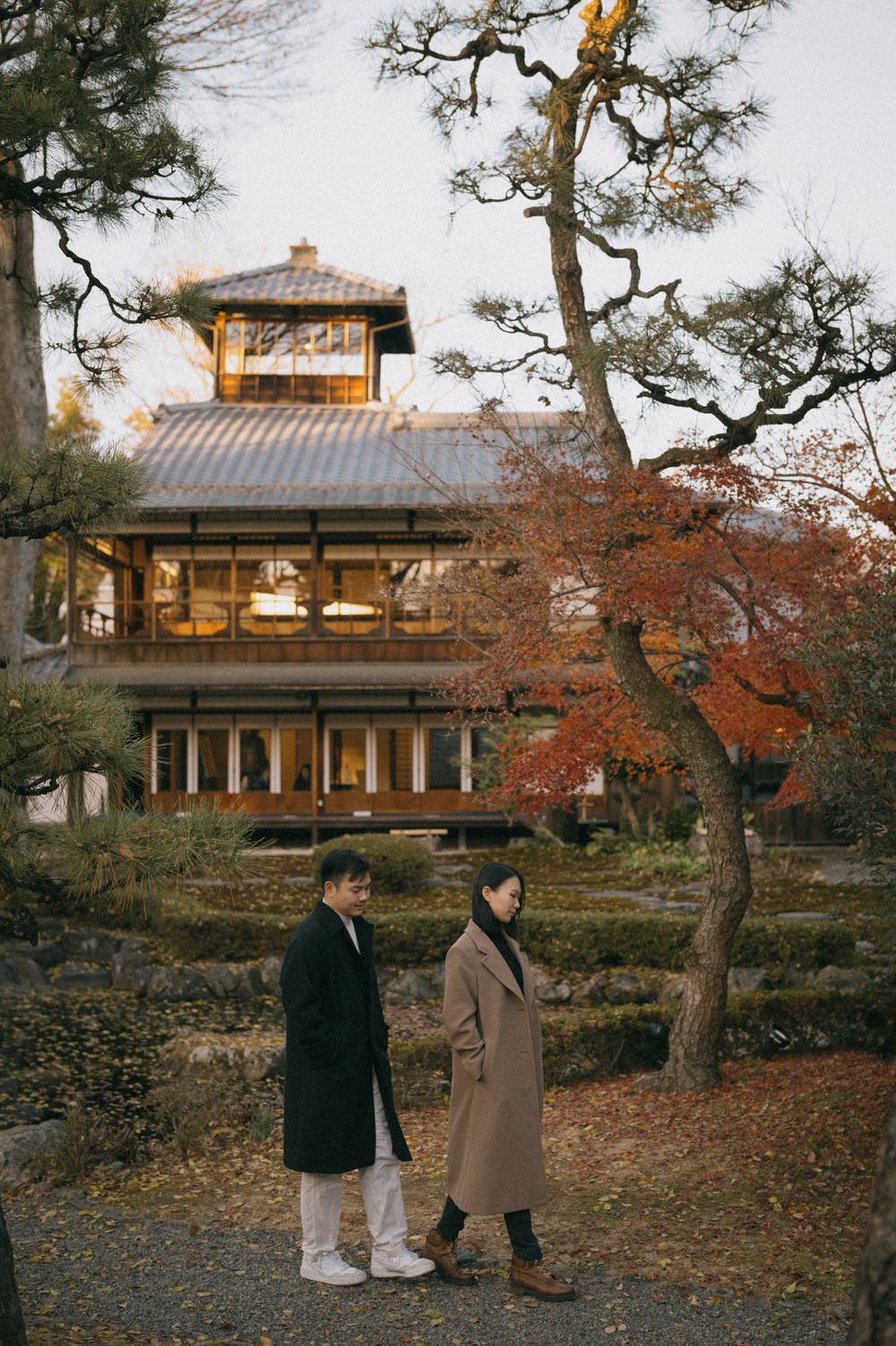 A couple walks hand in hand through a Japanese garden with autumn foliage and a traditional building.