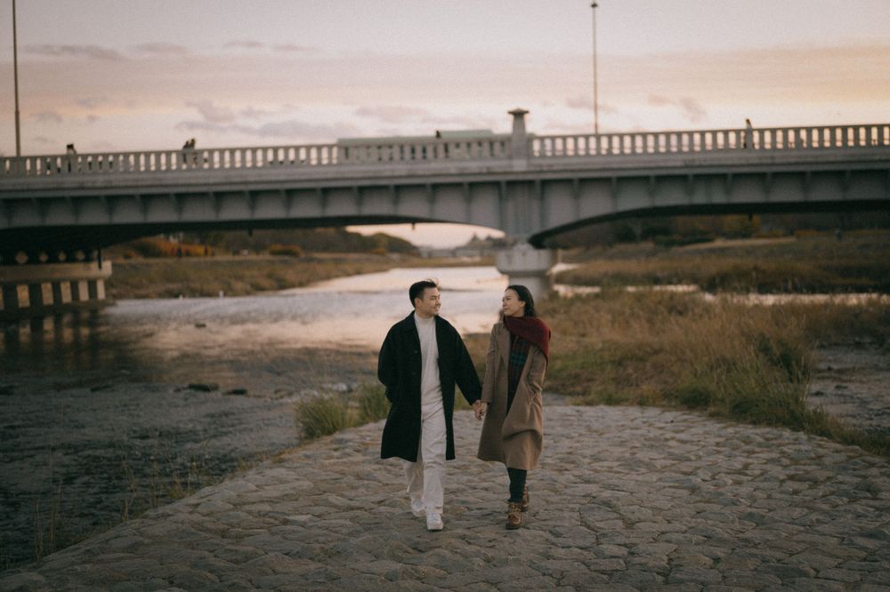 Couple holding hands and walking along a cobblestone riverside path near a bridge at sunset.