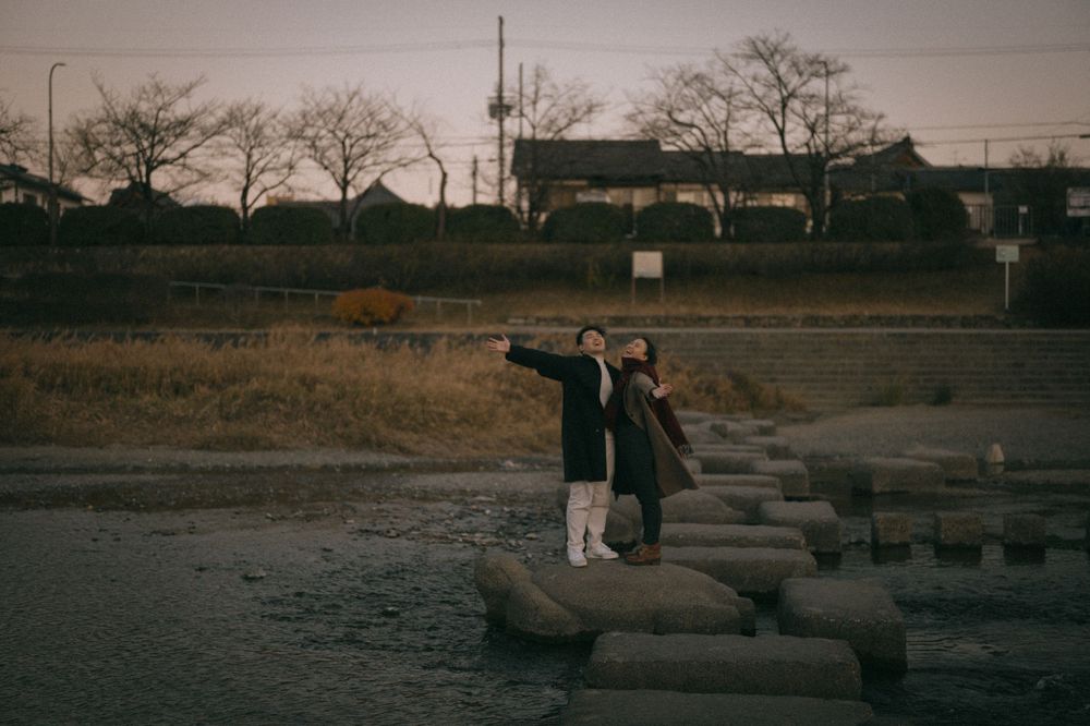 Couple standing on stone barriers by a riverbank pointing into the distance at dusk.