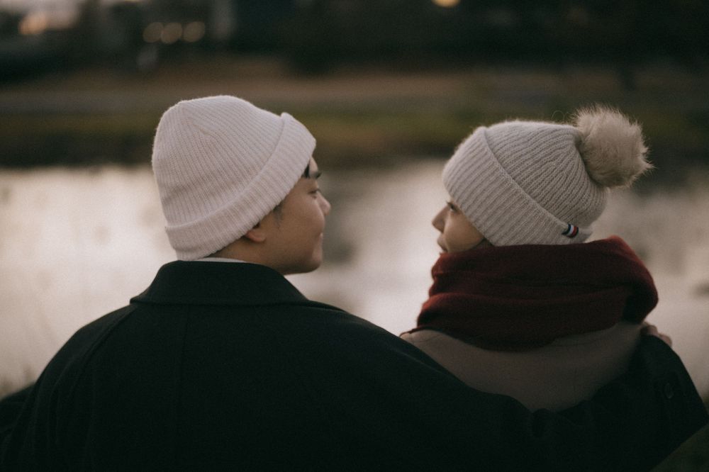 Couple wearing matching knit beanies gazing at each other by a reflective body of water.
