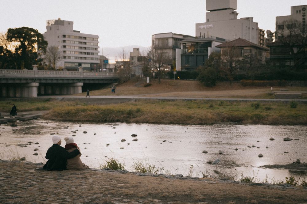 Couple sitting together on riverbank at dusk with city buildings reflected in calm water behind them.