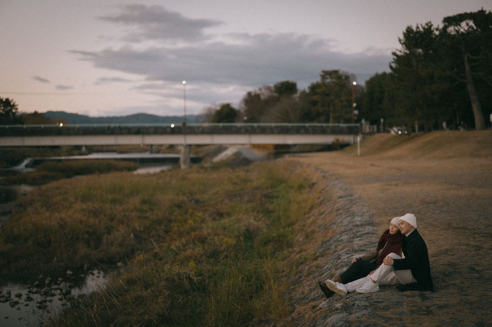 Couple sitting closely together on a dirt path at twilight near a serene waterway with mountains.