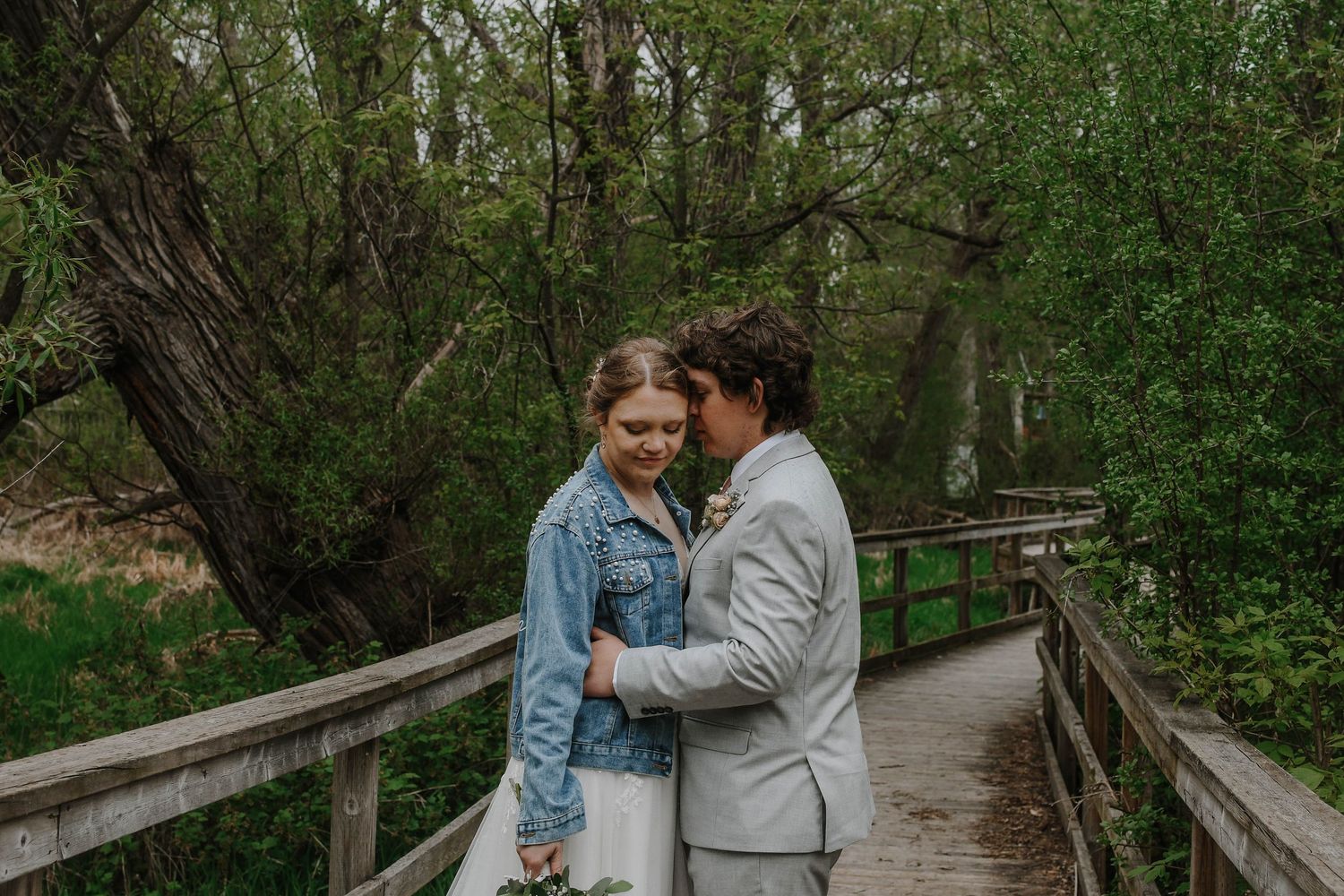 A romantic couple embraces on a wooden boardwalk surrounded by lush green forest scenery.