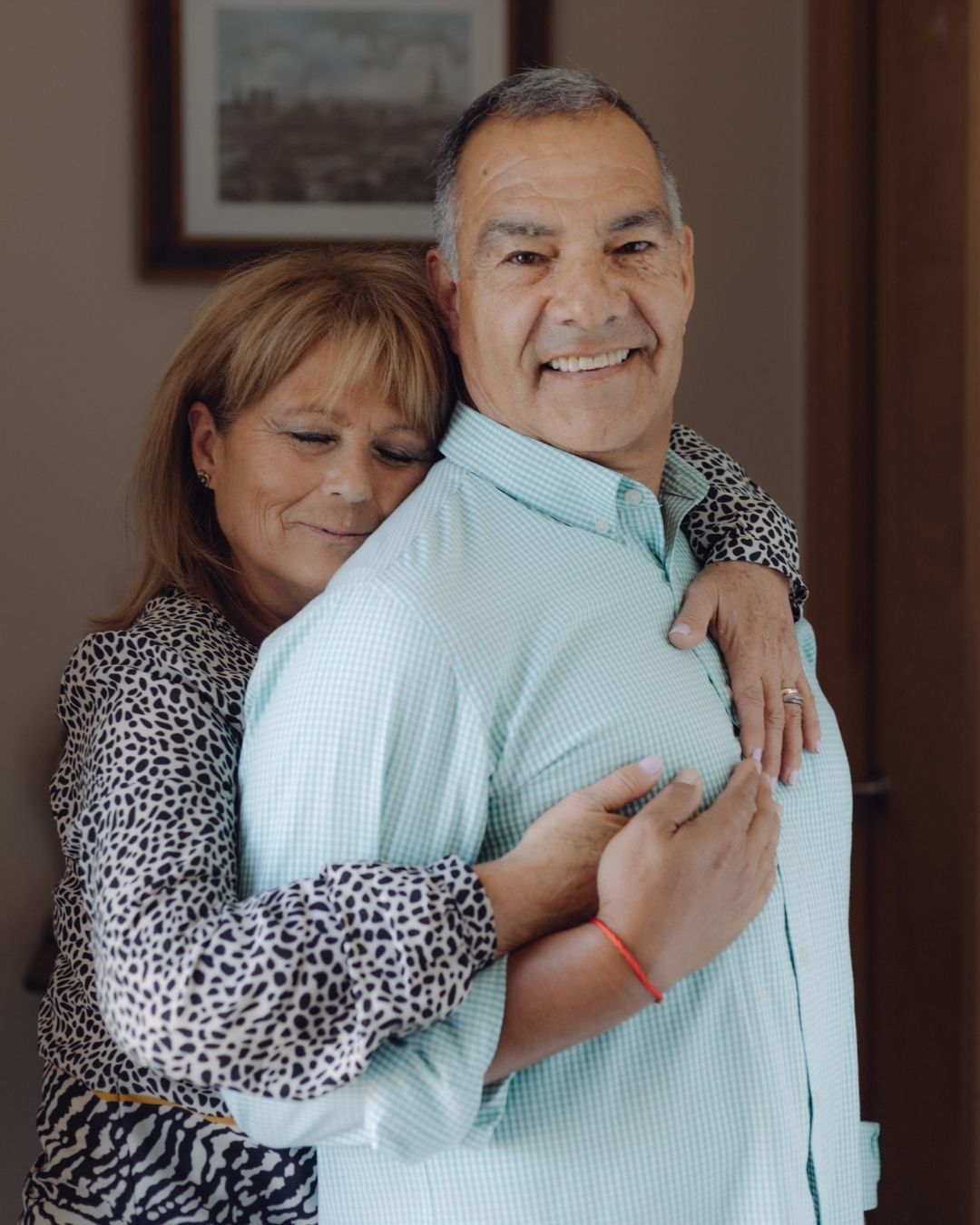 Mature woman hugging a smiling man from behind in a warm, affectionate indoor portrait.