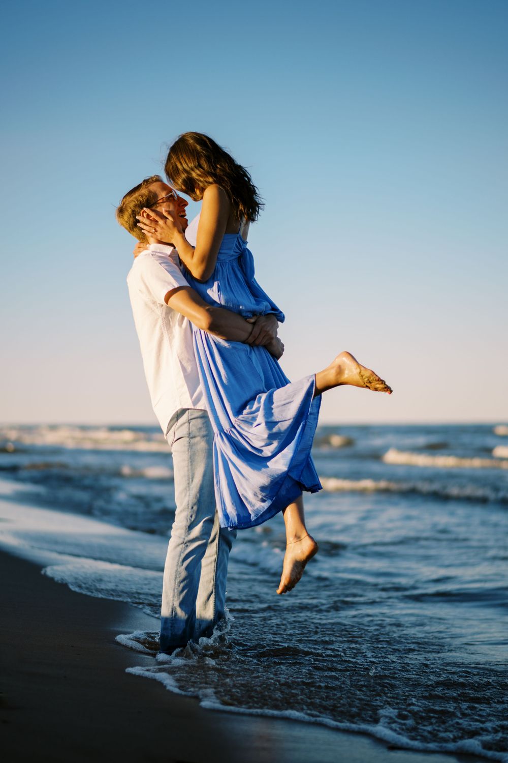 A couple in blue and white embrace on a beach at sunset while waves crash at their feet.