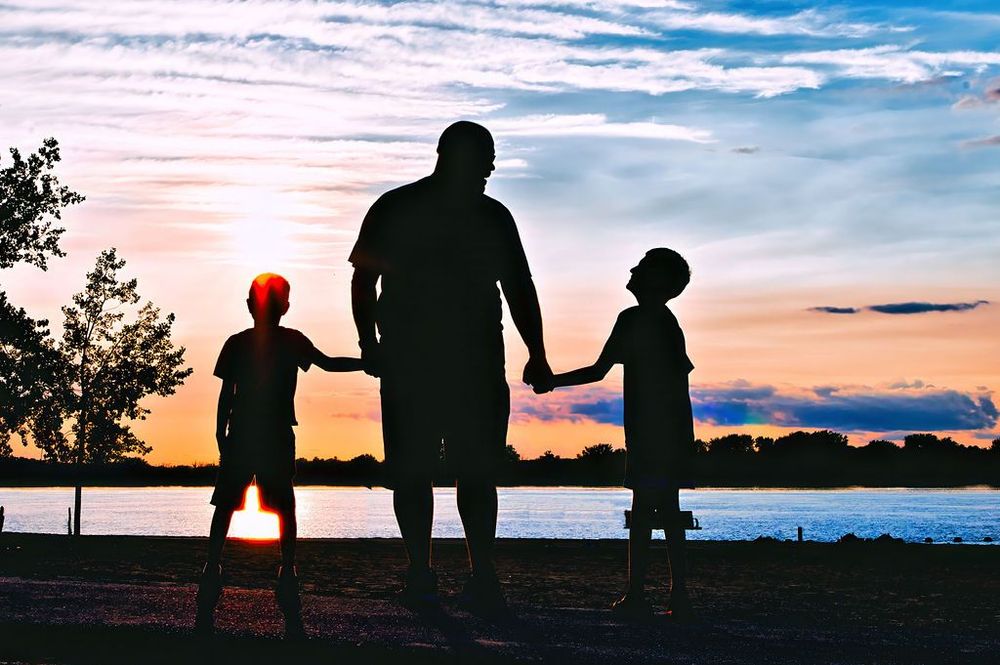 Silhouette of three people holding hands while watching a colorful sunset over a lake.