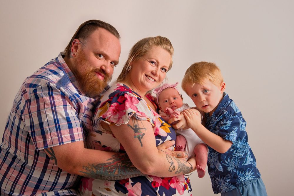 A family poses together in studio photography wearing casual clothing in pink and blue tones against neutral background.