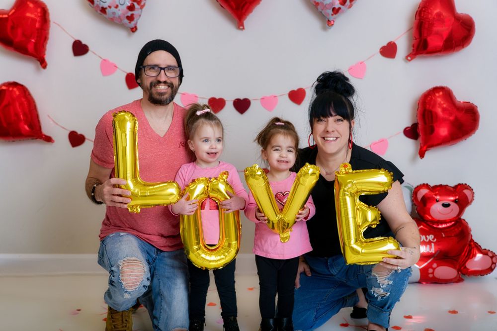 A family holds gold LOVE letters while posing against a white wall decorated with red heart balloons and valentines decor.