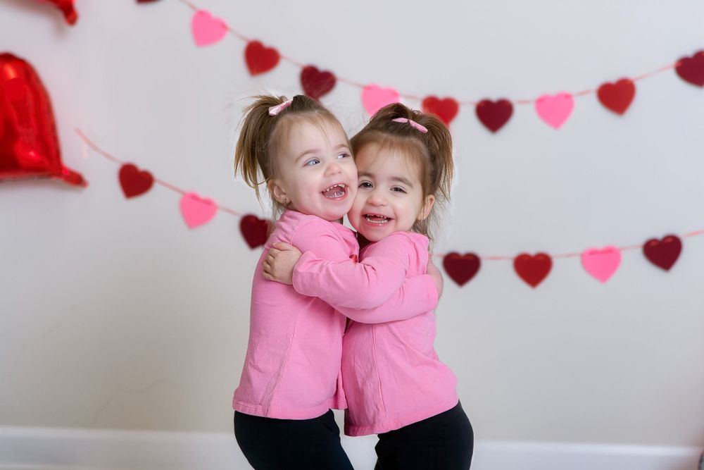 Two children in matching pink sweaters hug and laugh together against a white wall decorated with red and pink heart garland.