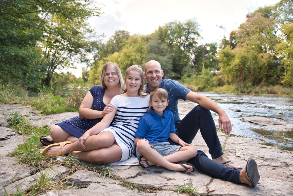 A family group sits together on rocky ground by a stream on a sunny autumn day.