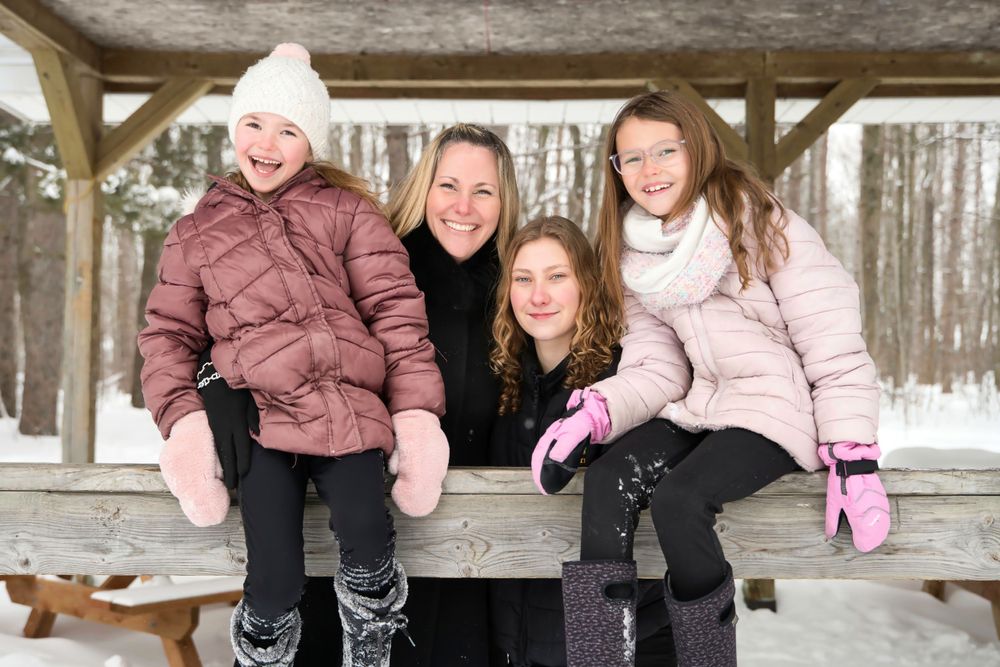 A winter scene with four people sitting together on a wooden bench in the snow wearing warm coats and winter accessories.