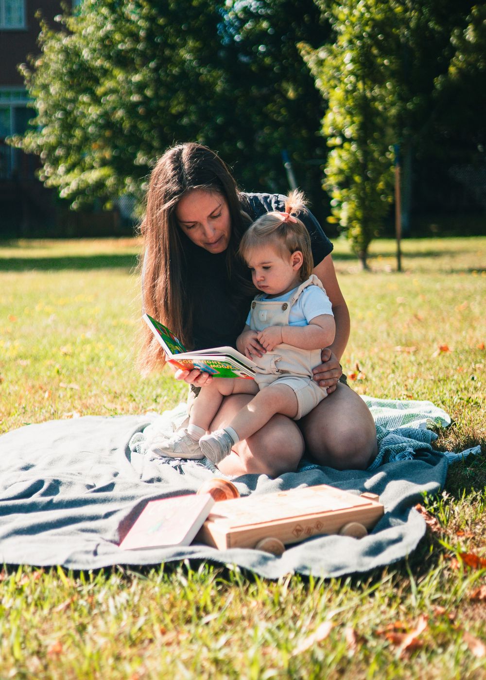 A parent and child share a quiet reading moment together on a blanket in a sunny park setting.