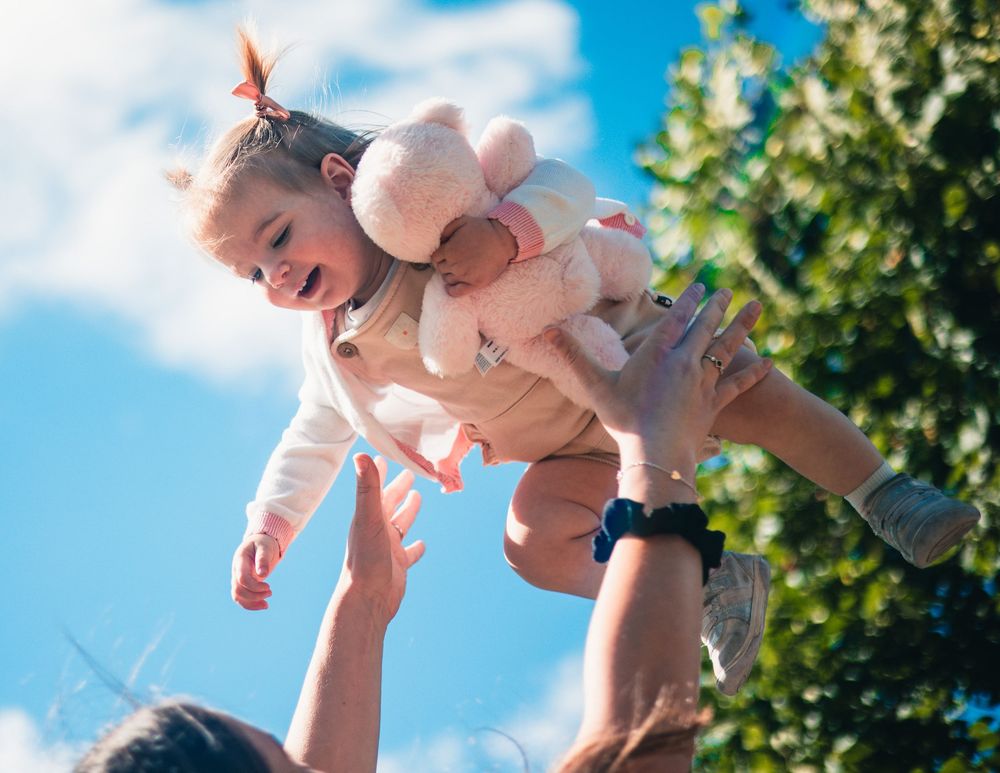 A baby wearing light colored clothing is lifted joyfully into the air against a bright blue summer sky with green trees.