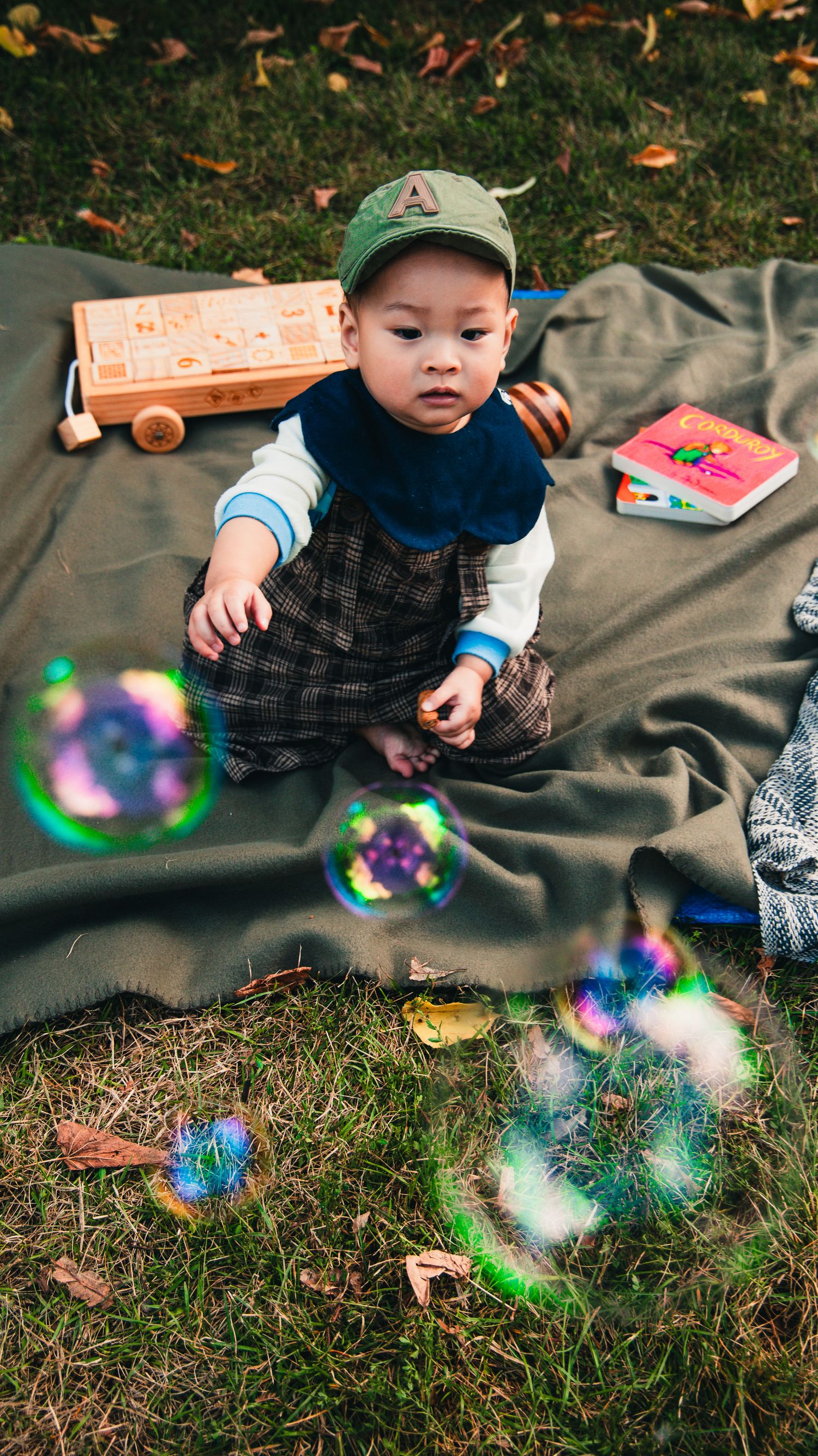 A small child on a blanket reaches for colorful soap bubbles floating in the air against a grassy background.