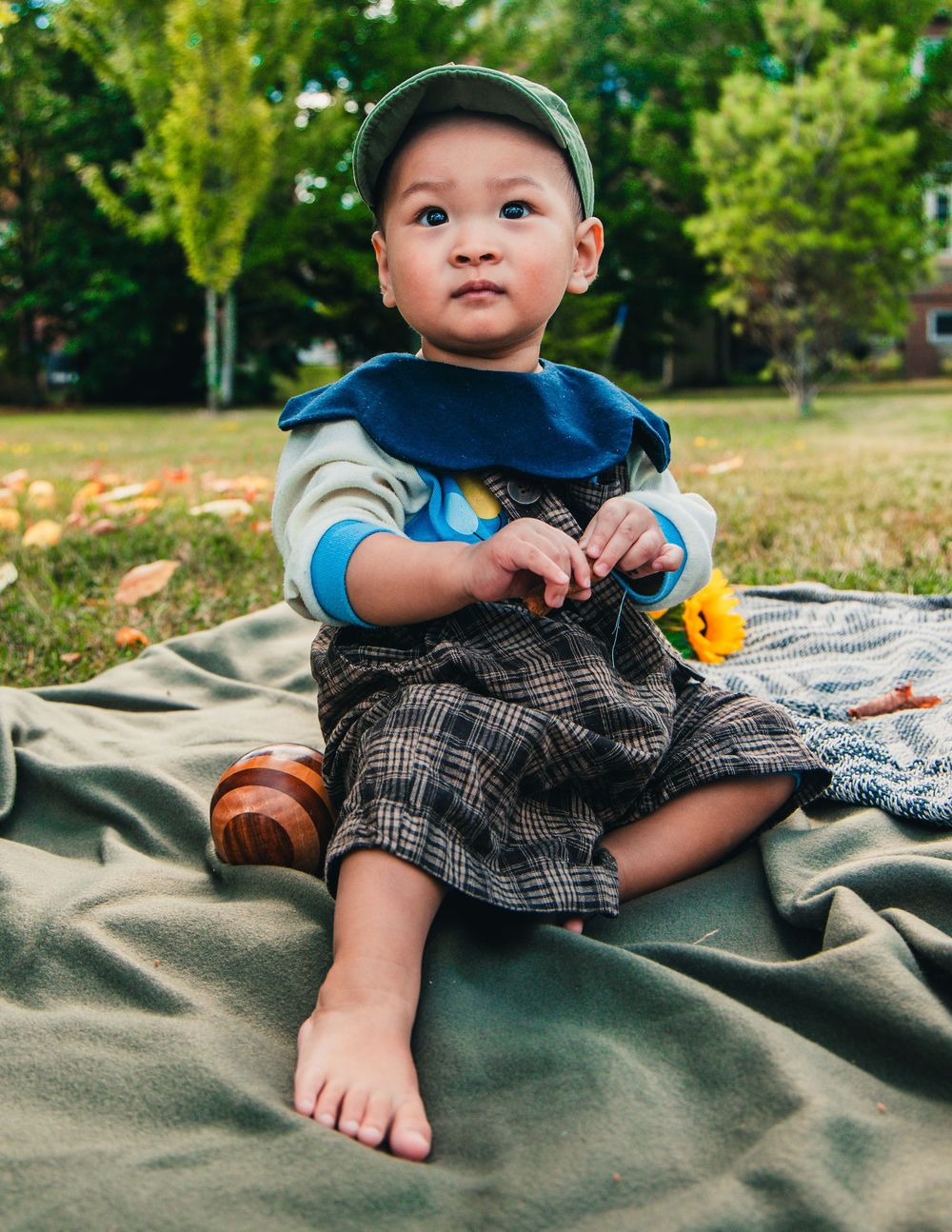 Small child wearing a baseball cap and plaid shorts sits on a blanket outdoors on a sunny day with greenery in background.
