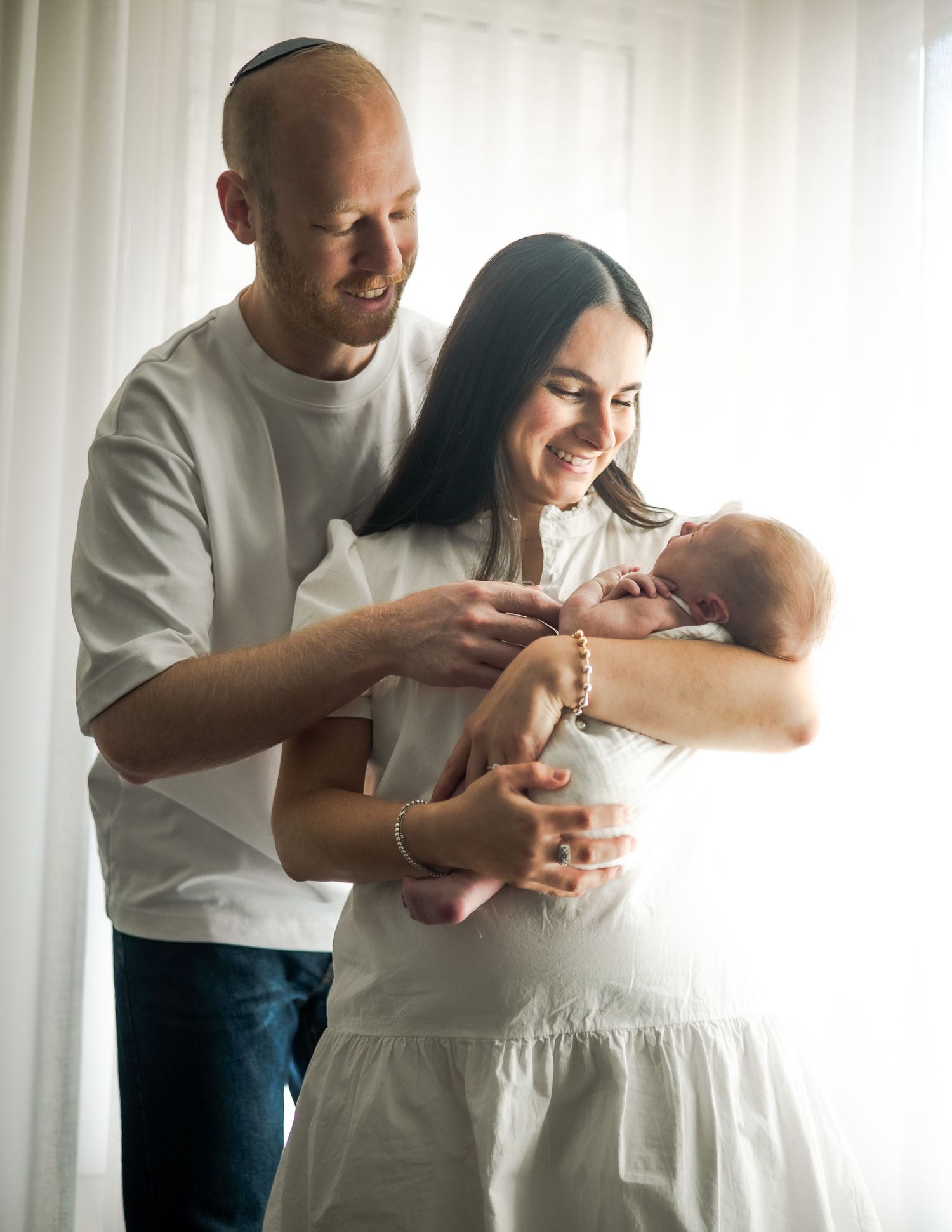 Three candid moments show a tender interaction between parent and baby in white clothing by a bright window.